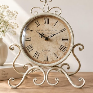Pode incluir: Um relógio de mesa de metal na cor creme com acabamento desgastado, apresentando um mostrador redondo com algarismos romanos pretos, pernas com volutas ornamentadas e uma alça decorativa. O mostrador exibe o texto Winston Clock Co. est. 1845 Quartz.
