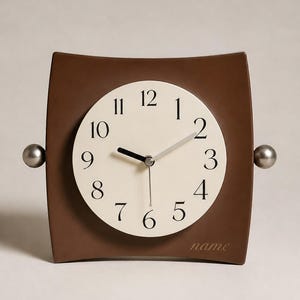 May include: A brown square-shaped clock with concave sides and a circular cream-coloured face. The clock features black Arabic numerals from 1 to 12, black hour and minute hands, a thin silver second hand, and the word name printed in the bottom right.