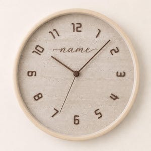 Puede incluir: Un reloj de pared redondo con marco de madera clara y esfera beige texturizada. El reloj presenta números de color marrón oscuro del 1 al 12, manecillas de color marrón oscuro y la palabra name escrita en cursiva debajo del número 12.