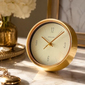 Pode incluir: Um relógio de mesa analógico redondo com uma moldura de metal dourado polido e um mostrador de cor creme. O relógio apresenta algarismos pretos nas posições das 12, 3, 6 e 9 horas, ponteiros dourados e a palavra "ENGLAND" impressa a preto.