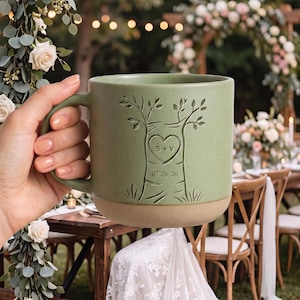 May include: A sage green ceramic mug with a speckled finish and a smooth tan base. The front features an embossed tree design with a heart carving containing the letters S+V and the date 07.26.26 engraved on the trunk of the tree.