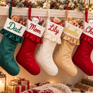 Puede incluir: Cinco calcetines navideños colgantes en verde, rojo, blanco, dorado y rojo. Cada uno tiene un puño blanco con borde de volantes y nombres bordados: Dad, Mom, Lily, Amelia y Chloe. Pequeños peluches y cajas de regalo asoman por las aberturas.