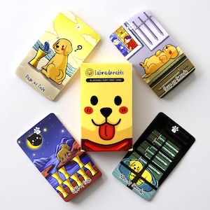 Può includere: Un mazzo di carte dei tarocchi Labradorable con una scatola gialla che mostra un muso di cucciolo e il testo 78 Adorable Puppy Tarot Cards. Quattro carte sono disposte attorno alla scatola, mostrando illustrazioni di cuccioli etichettate Page of Cups, Four of Swords, Eight of Cups e Nine of Swords.