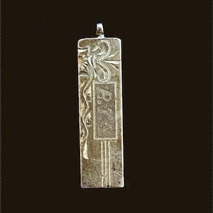 May include: A rectangular silver-toned metal pendant featuring an engraved floral design on the left side and the letters R.T.S. enclosed within a rectangular border on the right. The pendant includes a small metal loop at the top for attachment.
