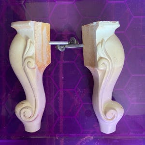 May include: Two off-white furniture legs featuring a curved, scroll-like design with carved detailing. Each leg includes a metal bolt and washer attachment extending from the side, designed for mounting to a base or frame of a piece of furniture.
