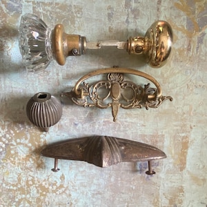 May include: A collection of hardware including a glass and brass door knob set, an ornate brass drawer pull with a swinging handle, a small round ribbed metal knob, and a wide, triangular-shaped metal drawer handle with two mounting screws.