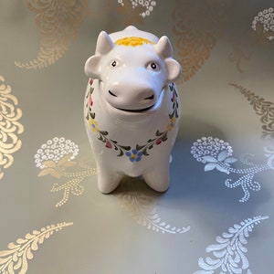 May include: White ceramic cow figurine with painted floral details in red, blue, and yellow. The cow has a yellow flower on its head and a smiling face.