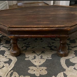 May include: A low-profile, octagonal wooden coffee table featuring a dark brown finish with visible wood grain. The table is supported by four thick, turned pedestal-style legs and has a flat, multi-sided top surface with a raised outer edge border.