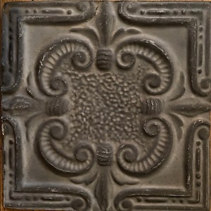 May include: A square metal ceiling tile featuring a dark gray finish with a distressed, weathered appearance. The surface displays a raised, symmetrical embossed pattern with central stippled texture, scrolling flourishes, and linear borders.