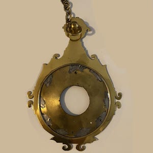 May include: A brass pendant with a circular opening and a decorative chain. The pendant has a worn, aged appearance.