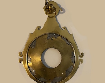 May include: A brass pendant with a circular opening and a decorative chain. The pendant has a worn, aged appearance.