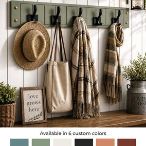 May include: A wall-mounted moss green coat rack with five black hooks holding a straw hat, a beige tote bag, a plaid blanket, and a plaid scarf. Below, a framed sign reads love grows here, alongside a wicker basket and a metal farm bucket.