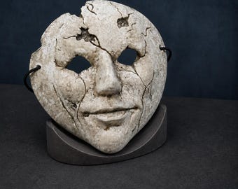 Könnte beinhalten: Eine verwitterte, cremeweiße Gesichtsmaske mit rissiger, abgenutzter Textur und tiefen Spalten über Stirn und Wangen. Die Maske hat zwei Augenöffnungen, eine ausgeprägte Nase und einen neutralen Mund sowie schwarze elastische Bänder an den Seiten.