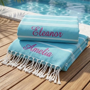 May include: Two light blue towels with white horizontal stripes and white fringe at the bottom edge. The top towel features the name Eleanor embroidered in pink script, while the bottom towel features the name Amelia embroidered in pink script.
