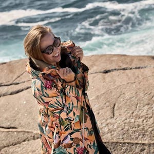 Peut inclure: Une veste à capuche couleur pêche ornée d'un imprimé botanique vibrant composé de feuilles vertes, de fleurs roses et de petits oiseaux. La veste comprend une fermeture éclair, un cordon de serrage à la taille et un petit écusson logo marron.