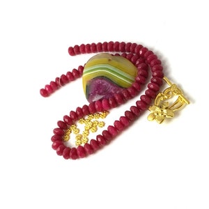 May include: A red gemstone bead necklace with a gold clasp and a large heart-shaped agate pendant with yellow, green, and purple stripes.