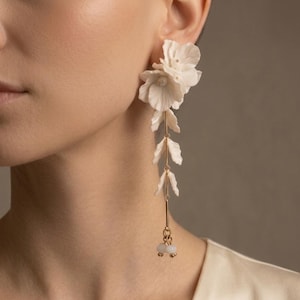 May include: A long drop earring featuring a cluster of white floral petals at the top, followed by a vertical gold-toned metal chain with three small white leaf-shaped accents, ending with two small round white beads at the bottom.