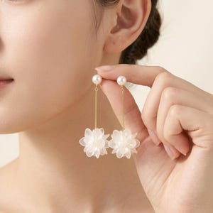 以下が含まれることがあります： 上部に小さな白いパールのスタッドが付いたゴールドトーンのドロップピアス。細いゴールドチェーンで、半透明の層状になった白い花形のペンダントにつながっています。繊細な揺れるデザインで、花のアクセントが特徴的です。