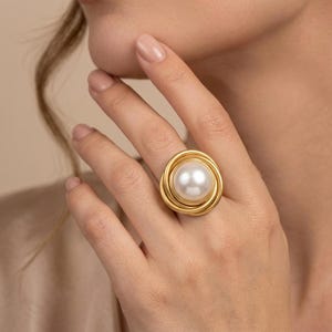 May include: A gold-toned ring featuring a central, smooth, round white pearl. The band is constructed from multiple overlapping, circular wire loops that create a layered, coiled effect around the pearl, providing a textured, metallic finish.