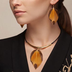 Op de afbeelding: Een sieradenset bestaande uit een ketting en hangende oorbellen met natuurlijke veren. De veren zijn bruin met een goudgele centrale streep. De ketting heeft een zwart koord met een gebogen goudkleurige metalen staaf die de veerhanger vasthoudt.