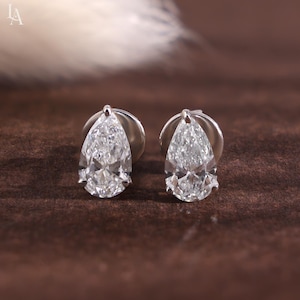 Puede incluir: Un par de pendientes de botón con piedras transparentes en talla pera montadas en garras de metal plateado. Cada pendiente tiene una parte superior puntiaguda y una base redondeada, mostrando una superficie facetada que refleja la luz.