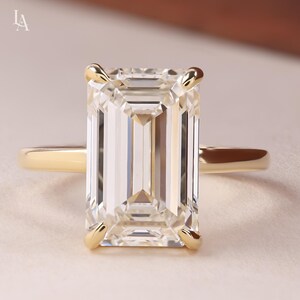 May include: A gold ring featuring a large, rectangular emerald-cut clear gemstone held by four prongs. The band is smooth and polished, showcasing a simple design that highlights the faceted stone. The setting is yellow gold, providing a classic contrast to the brilliant, transparent center stone.