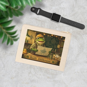 Op de afbeelding: Een beige etui met rits en een bedrukte illustratie van een kikker met een veiligheidshelm en geruit overhemd, die een blauwdruk vasthoudt. De tekst 'Built Different' staat onderaan. Een zwarte smartwatch ligt ernaast.