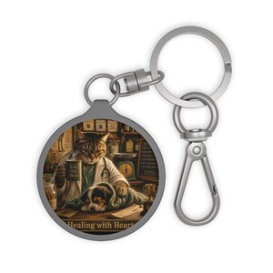 May include: A round silver keychain featuring a graphic of a tabby cat veterinarian wearing a white coat and stethoscope, holding a mug labeled Cat Vet Hero while tending to a puppy. The text Healing with Heart is printed at the bottom.