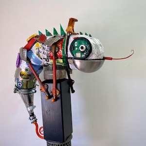May include: A sculpture of a creature constructed from recycled materials including circuit boards, newspaper, metal components, and a silver bowl. The figure features a long red wire tongue, green circuit board spikes, and a black rectangular base.