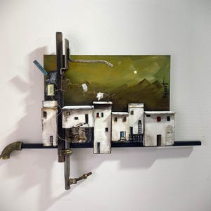 May include: A mixed-media wall sculpture featuring a painted olive-green landscape background with mountain silhouettes. Attached are several small, white, block-like structures resembling houses, accented with metal pipes and wire elements.