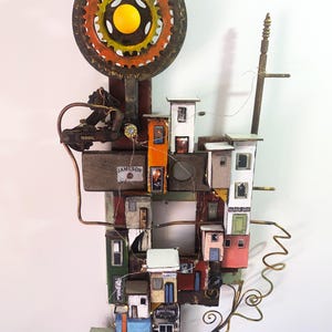 May include: A wall-mounted assemblage art piece featuring a large metal gear with a yellow center, connected to a cluster of miniature wooden buildings. The structures include signs for a bakery and barber shop, with copper wire accents and a Jameson label.