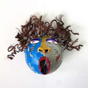 May include: A spherical papier-mâché sculpture of a face with brown yarn hair. The face is split into blue and gray sections with a yellow nose, orange-rimmed eyes, and a wide, open red mouth with a black interior. Text fragments are visible.