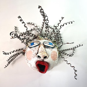 May include: A wall-mounted sculpture of a human face with a textured white surface, bright blue eyes, pink cheeks, and an open red mouth. The head is adorned with numerous metallic, silver-colored spiral curls extending outward in all directions.