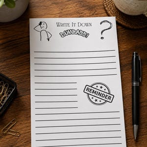 May include: A white rectangular notepad with black horizontal lines. The top features the text "WRITE IT DOWN DUMBASS!" alongside a stick figure and a question mark. A circular stamp graphic with the word "REMINDER" and stars is printed on the lower right.