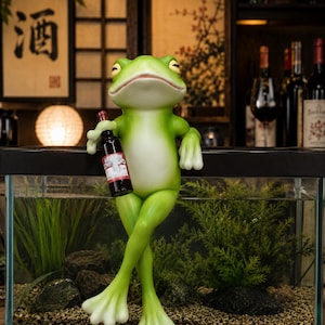 May include: A green frog figurine with a light-colored belly, sitting with legs crossed on the edge of a glass aquarium. The frog holds a small dark bottle with a red label in its right hand. The frog has a smooth texture and closed eyes.