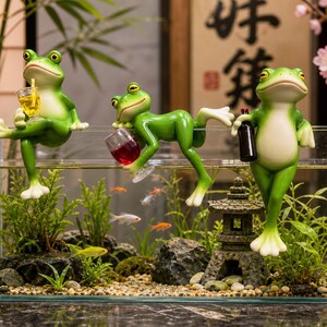 May include: A rectangular glass aquarium containing water, green aquatic plants, small fish, and a stone pagoda ornament. Three green frog figurines are perched on the top edge of the tank, holding a yellow drink, a red drink, and a dark bottle.