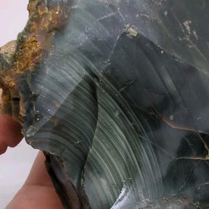 May include: A raw, dark green and black obsidian stone featuring a smooth, glassy surface with visible conchoidal fractures and subtle, light-colored linear striations running across the polished-looking face, with rough, earthy brown edges.