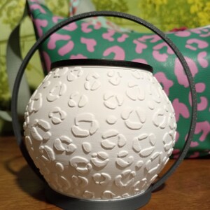 May include: A white spherical wax warmer featuring a raised leopard print texture. The warmer is framed by a thin, dark gray circular metal handle that extends from the base, creating a distinct silhouette for this functional home accessory.