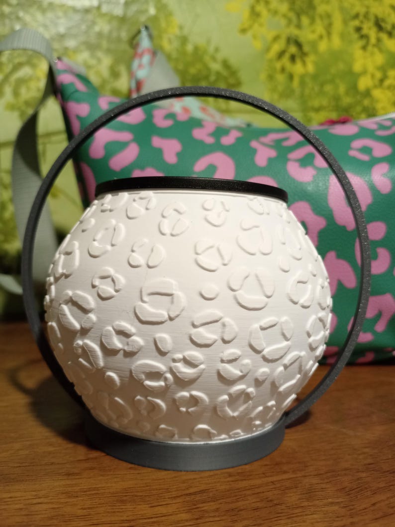 May include: A white spherical wax warmer featuring a raised leopard print texture. The warmer is framed by a thin, dark gray circular metal handle that extends from the base, creating a distinct silhouette for this functional home accessory.