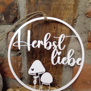 Könnte beinhalten: Ein weißer, kreisförmiger Wandschmuck mit dem deutschen Schriftzug "Herbst liebe" in Schreibschrift. Unter dem Text befinden sich zwei stilisierte Pilzillustrationen, wobei der größere Pilz ein kleines Herzdetail und eine Schleife am Stiel aufweist.