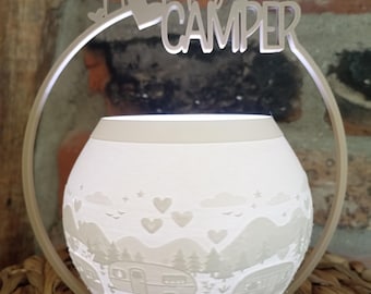 May include: A white, spherical, illuminated lamp featuring a relief design of vintage campers, pine trees, mountains, and hearts. A circular frame surrounds the lamp, topped with a cutout sign that reads Happy CAMPER in a script and block font.