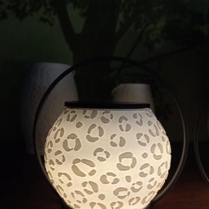May include: A spherical wax warmer featuring a white, translucent body with a cut-out leopard print pattern. The device is illuminated from within, casting a soft glow. It is framed by a thin, black circular metal handle and sits on a black base.