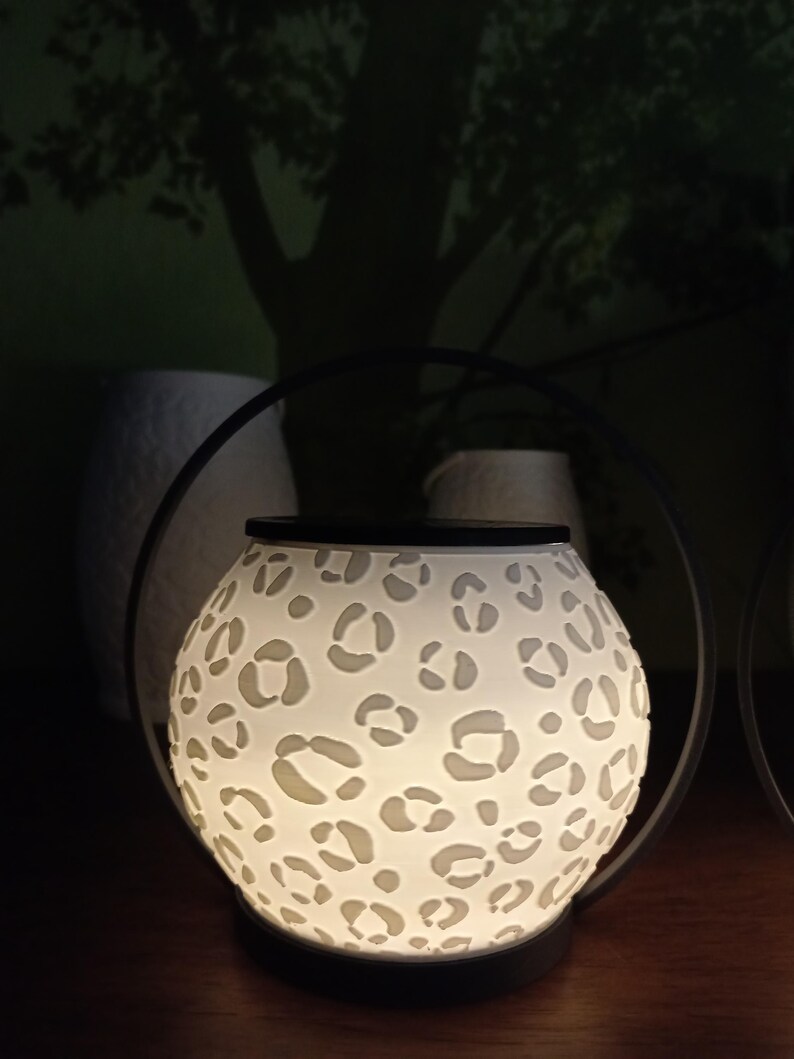 May include: A spherical wax warmer featuring a white, translucent body with a cut-out leopard print pattern. The device is illuminated from within, casting a soft glow. It is framed by a thin, black circular metal handle and sits on a black base.