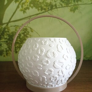 May include: A white spherical planter featuring a raised leopard print texture. The planter is set within a tan circular frame that acts as both a base and a handle, creating a modern and structured appearance for this decorative plant container.