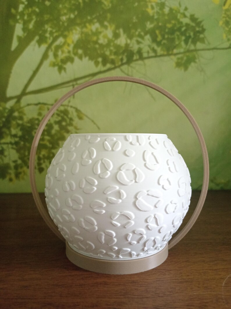 May include: A white spherical planter featuring a raised leopard print texture. The planter is set within a tan circular frame that acts as both a base and a handle, creating a modern and structured appearance for this decorative plant container.