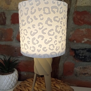 Könnte beinhalten: Eine Tischlampe mit zylindrischem Lampenschirm, der ein weißes und hellgraues Leopardenmuster aufweist. Die Lampe hat einen beigen, gedrehten Stativfuß. Der Schirm wird von innen beleuchtet und wirft ein sanftes Licht durch das Muster.