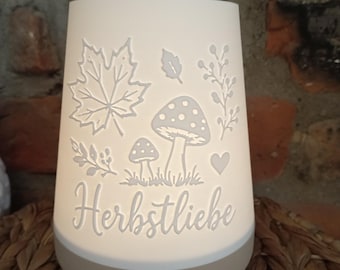May include: A white, conical, illuminated lamp featuring etched designs of a maple leaf, mushrooms, a small heart, and sprigs. The base is a solid, opaque light beige color. The word "Herbstliebe" is inscribed in a cursive script on the front.