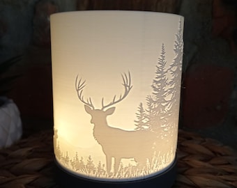 May include: A cylindrical white lamp shade featuring a backlit silhouette of a deer standing in a forest with pine trees. The shade is mounted on a solid dark circular base, emitting a warm glow that highlights the intricate cut-out design.