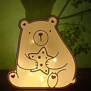 Könnte beinhalten: Eine leuchtende, cremeweiße, bärenförmige Lampe mit dunkler Umrandung. Der Bär sitzt und hält einen Stern mit kleinen sternförmigen Ausstanzungen. Die Lampe strahlt ein warmes Licht aus und betont die herzförmigen Details an den Ohren und Füßen.