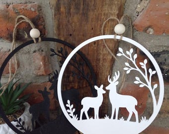 May include: Two circular hanging ornaments, one black and one white, each featuring a laser-cut silhouette of two deer standing in a forest scene with a tree branch. Each ornament includes a natural jute twine loop and a small wooden bead.